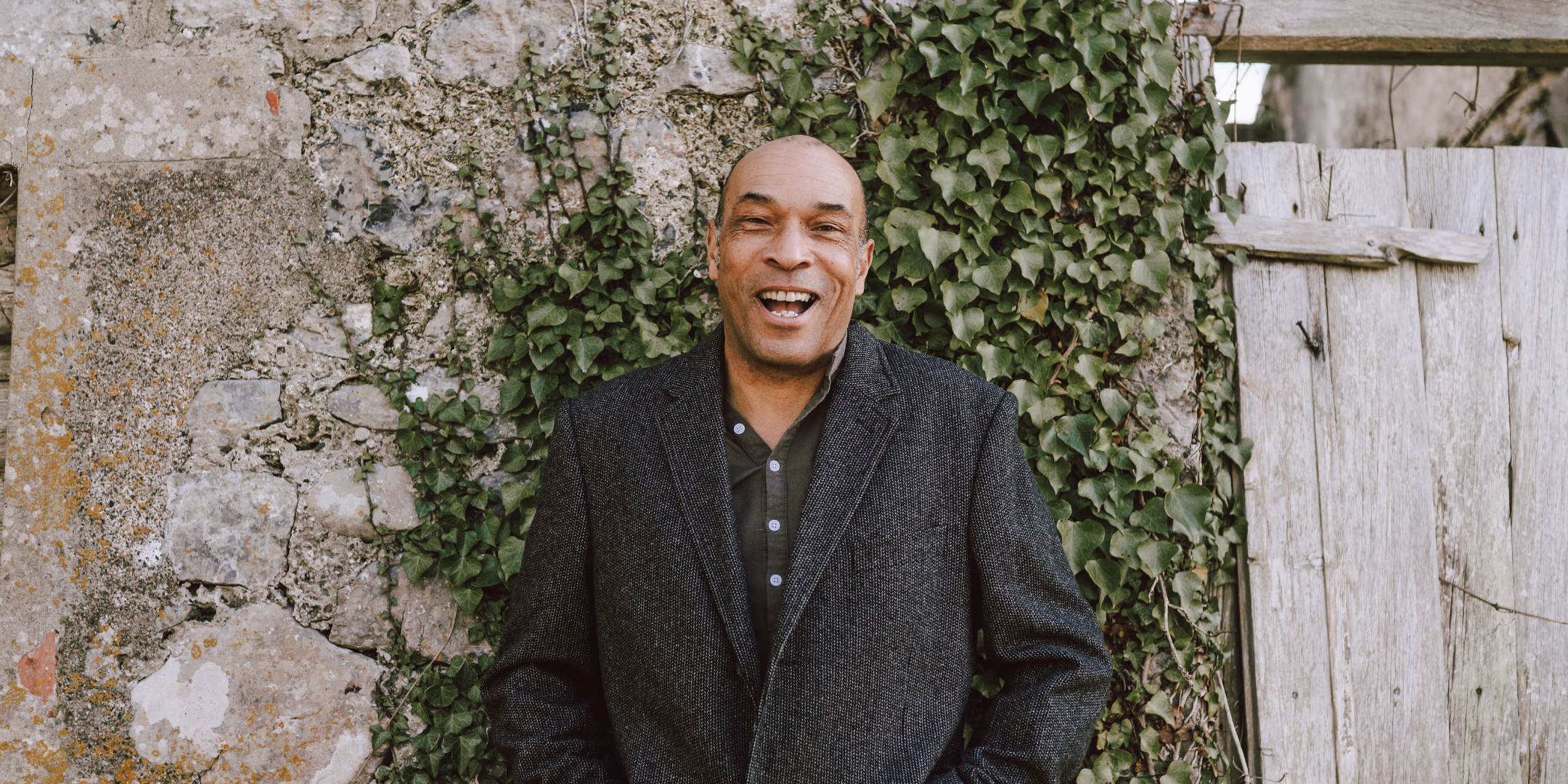 Phil Okwedy is standing in front a wall wearing a dark grey jacket and green top. The wall has some climbing ivy in the background and an old door to the right.