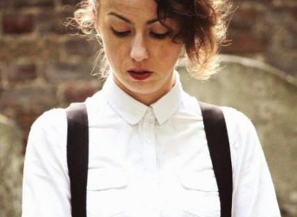 a white woman with long brown hair tied back, wearing a white shirt and black braces