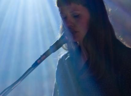 a white woman with long dark hair singing into a microphone on stage. there is a strong shaft of light.