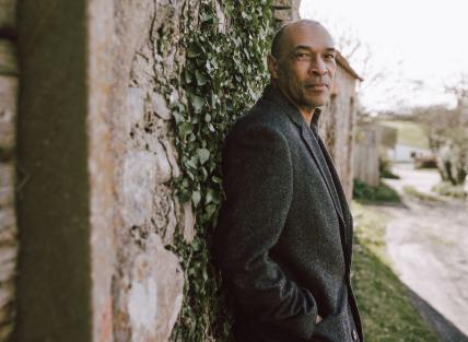 a black man with cropped hair, wearing a winter coat and leaning against a wall